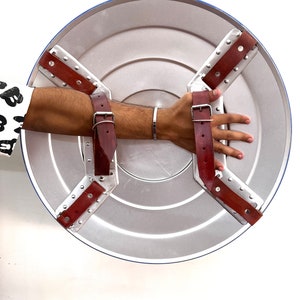 Puede incluir: Un escudo plateado con borde azul y correas de cuero marr&oacute;n. El escudo presenta un dise&ntilde;o circular con anillos conc&eacute;ntricos. Las correas est&aacute;n remachadas al escudo y sujetas con hebillas met&aacute;licas. Un accesorio cl&aacute;sico de superh&eacute;roe.