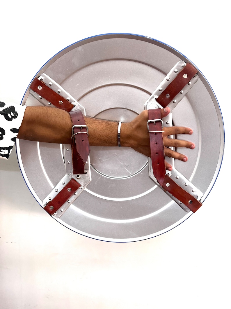 Puede incluir: Un escudo plateado con borde azul y correas de cuero marr&oacute;n. El escudo presenta un dise&ntilde;o circular con anillos conc&eacute;ntricos. Las correas est&aacute;n remachadas al escudo y sujetas con hebillas met&aacute;licas. Un accesorio cl&aacute;sico de superh&eacute;roe.