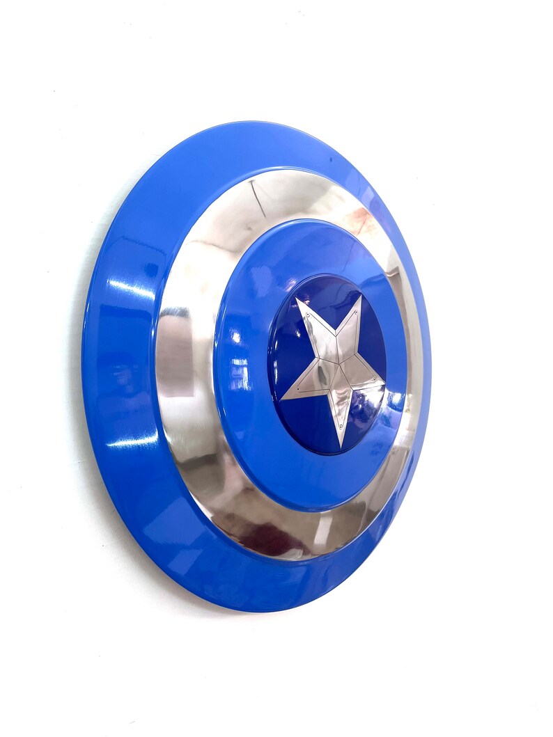 Puede incluir: Un escudo azul y plateado con una estrella plateada en el centro. El escudo tiene un dise&ntilde;o circular con anillos conc&eacute;ntricos azules y plateados. Es un accesorio o art&iacute;culo de colecci&oacute;n.