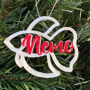 Peut inclure: Ornement de Noël blanc en forme de poisson avec le nom "Nemo" en script rouge. L'ornement est suspendu à une ficelle argentée sur un fond de branches vertes de sapin de Noël.
