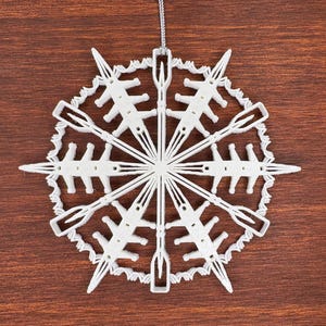 May include: A white, 3D-printed snowflake ornament with a complex, geometric design featuring pointed, arrow-like arms and a textured, scalloped outer ring. The ornament is suspended by a thin, silver-colored string attached to the top point.