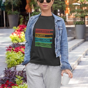 May include: A person wearing a black t-shirt with the words "Diversity, Equity, Inclusion, Belonging" printed in rainbow colors. The person is wearing a denim jacket and grey sweatpants.