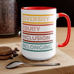 May include: White ceramic mug with a red rim and handle. The mug has the words "Diversity", "Equity", "Inclusion", and "Belonging" printed in a retro style with colorful stripes.