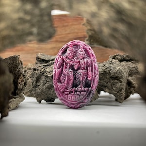 Könnte beinhalten: Ein geschnitzter Anhänger aus rosa Edelstein mit einer detaillierten Darstellung von zwei Figuren in sitzender Pose. Der Anhänger ist oval geformt und hat eine glatte, polierte Oberfläche.