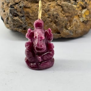 May include: A carved, deep red pendant in the shape of an elephant-headed deity, with a gold-coloured loop for hanging. The pendant is set against a neutral background, highlighting the intricate details of the sculpture.