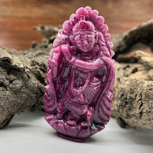 Könnte beinhalten: Ein geschnitzter Anhänger aus rosafarbenem Rubin, der eine Gottheit mit filigranen Details darstellt. Der Anhänger ist vor einem rustikalen Holzhintergrund platziert.