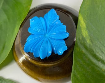 May include: A bright blue flower-shaped brooch with a carved design. The brooch is sitting on a brown wooden surface.