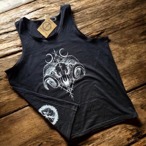 Könnte beinhalten: Ein schwarzes ärmelloses Tanktop mit einem weißen Grafikdruck eines Widder-Schädels mit geschwungenen Hörnern, geometrischen Linien und Mondsicheln. Ein kreisförmiges Logo ist am unteren Saum aufgedruckt und ein braunes Papieretikett ist am Halsausschnitt befestigt.