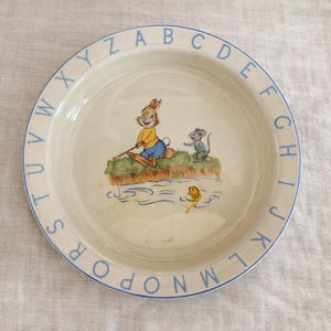 May include: A round, off-white ceramic plate with a blue rim and alphabet letters. The plate features a cartoon illustration of a rabbit fishing with a mouse and a fish. The plate is likely for children.