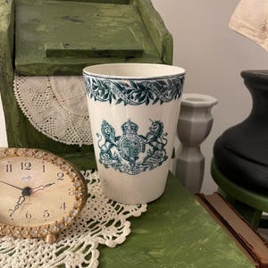 May include: A white ceramic tumbler with a teal and white crest design. The tumbler features a detailed coat of arms with lions and a crown, and is decorated with a floral border. The tumbler is displayed on a green surface.