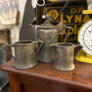 May include: Antique silver-toned tea set, including a teapot and two small pitchers. The set is displayed on a wooden surface, with a yellow scale and vintage items in the background. The tea set has ornate handles and a decorative finial.