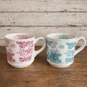 May include: Two white ceramic mugs with handles, each featuring a pastoral scene in a toile design. One mug has a red and white design, the other has a teal and white design. The text "Catching the Mail" is printed on the bottom of each mug.
