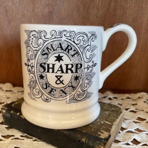 May include: White ceramic mug with a curved handle, featuring the words "SMART, SHARP & SEXY" in black script within an ornate oval design. The mug sits on a vintage book, showcasing a classic, elegant style.