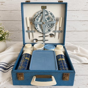 May include: Vintage blue picnic basket with a white handle, containing plates, cutlery, cups, and thermoses. The plates have a blue and white floral design. The thermoses have a blue and gold plaid pattern. The basket is open, revealing its contents.
