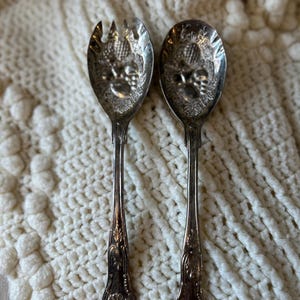 May include: A pair of vintage silver serving utensils, a spoon and a fork, with ornate floral and fruit designs on the bowls. The handles have a detailed, decorative pattern. The utensils rest on a cream-colored, textured surface.