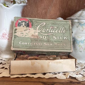 May include: Vintage Corticelli Spool Silk box, open to reveal spools of thread. The box is light green with a cat illustration and text that reads "Corticelli Spool Silk". The box is worn, suggesting age. A charming piece for sewing enthusiasts.