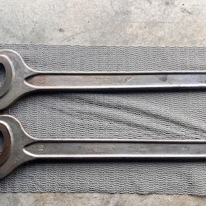 Peut inclure: Deux clés à molette anciennes en acier, de couleur métallique foncée, avec un design incurvé. Elles sont posées sur une surface grise texturée. Chaque clé a un trou à l'extrémité du manche.