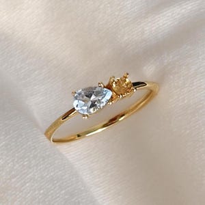 May include: A gold ring with a pear-shaped, pale blue gemstone and a smaller, round, yellow gemstone. The ring has a thin gold band, and the gemstones are set close together. The ring is shown on a white, textured surface.