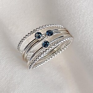 May include: A multi-band ring featuring a combination of smooth silver bands and twisted rope-textured silver bands. Three small, round, dark blue stones are set into the bands, creating a layered appearance with a polished metallic finish.
