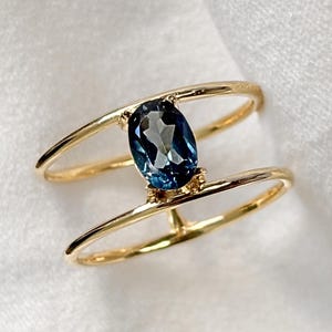 May include: A gold ring with a large, oval-cut, deep blue gemstone. The ring's design includes two parallel bands that curve around the finger, giving it a modern, open appearance. The gemstone is the central feature of the ring.