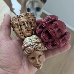 Peut inclure: Une main tient trois petites sculptures de visages humains. L'une est dorée et divisée en trois visages, une autre est un buste doré et la troisième est un visage rouge hurlant. Un chien est en arrière-plan.