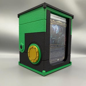 Könnte beinhalten: Eine rechteckige, schwarz-grüne Deck-Box mit strukturierter Oberfläche, einem goldenen, kreisförmigen Akzent an der Seite und einem transparenten Sichtfenster auf der Vorderseite, in dem eine Doctor Doom, Ring of Latveria Karte steckt.