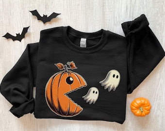 May include: Black sweatshirt featuring a Halloween-themed graphic. The design includes an orange pumpkin with a bite taken out of it, eating two white ghosts. Two black paper bats are above the sweatshirt, and a small orange pumpkin is on the right.