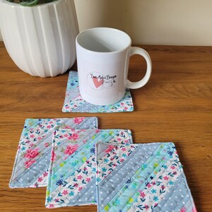 May include: Four blue and white quilted fabric coasters with a floral pattern. The coasters are arranged in a stack with the top coaster slightly offset. A white mug with the text "Dani Makes Designs" is sitting on top of the coasters.