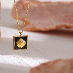 May include: A gold chain necklace with a square pendant featuring a black background and a gold seashell design.