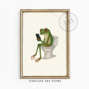 Può includere: Una stampa artistica incorniciata raffigurante una rana verde con occhi arancioni seduta su un water bianco mentre guarda uno smartphone nero. L'opera è racchiusa in una cornice color oro metallizzato con il testo TIMELESS ART STORE.
