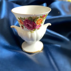 Può includere: Un vaso in porcellana bianca con un motivo floreale in tonalità rosa, giallo e verde. Il vaso ha due manici e un bordo dorato attorno al bordo.