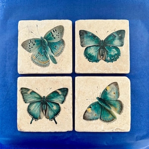 Pode incluir: Conjunto de quatro  tapetes quadrados com um design de borboleta azul. Cada tapete tem uma borboleta diferente sobre um fundo branco.
