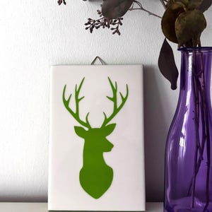 Könnte beinhalten: Ein weißes rechteckiges Wandbild mit einer grünen Hirschkopf-Silhouette. In der Nähe befindet sich eine lila Vase mit getrockneten Zweigen und Blättern. Das Kunstwerk wird mit einem Metallhaken aufgehängt. Die Vase ist etwa 25 cm hoch.