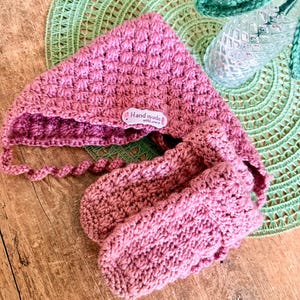 Könnte beinhalten: Handgemachtes Set aus altrosa Häkelmütze und passenden Fäustlingen. Die Mütze hat ein strukturiertes, sternförmiges Muster und Bindebänder. Die Fäustlinge sind ebenfalls mit dem gleichen Muster gehäkelt. Ein kleines Etikett auf der Mütze lautet "Hand made with love".