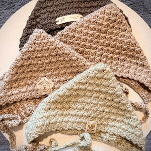 Könnte beinhalten: Vier handgefertigte Babyhauben aus Häkelarbeit in Braun- und Beigetönen. Jede Haube hat ein strukturiertes Stichmuster und Bindebänder. Die Hauben haben kleine Etiketten mit der Aufschrift "Made with Love" und "handmade with love".