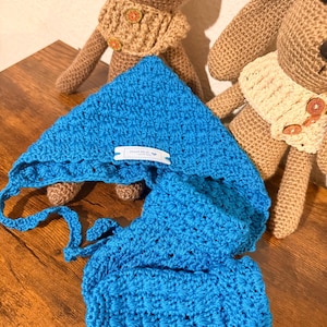 Könnte beinhalten: Ein leuchtend blaues Häkel-Baby-Set mit Mütze und Schal. Die Mütze hat Bindebänder und ein kleines weißes Etikett. Im Hintergrund sind zwei gehäkelte Tierpuppen, eine mit beigem Kragen und die andere mit cremefarbenem Kragen.