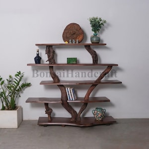 May include: A wooden shelf unit with a unique tree branch design. The shelves are made of dark brown wood and the branches are natural in color. The shelf unit has five shelves and is decorated with a small wooden circle, a green box, a blue bottle, a green vase with a plant, and a blue and white patterned vase.
