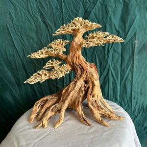 May include: A wooden bonsai tree sculpture with intricate detailing. The sculpture has a light beige colour, with a natural wood grain. The tree's trunk and branches are visible. The background is a green fabric.