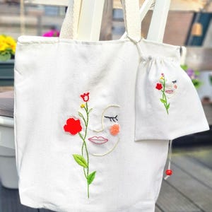 Op de afbeelding: Witte canvas tote bag en bijpassende trekkoordtas, beide geborduurd met een bloemenontwerp en een gestileerd gezicht. De tote bag heeft een grote rode bloem en groene stengel. De tas heeft een kleinere versie van hetzelfde ontwerp.