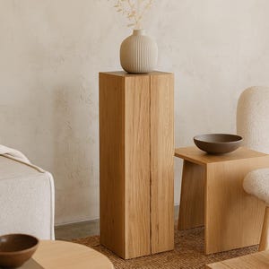 May include: A wooden pedestal with a vase and dried plant arrangement. A small, square wooden side table with a dark brown bowl is next to the pedestal. A woven wall hanging is in the background. The pedestal is approximately 100 cm tall.