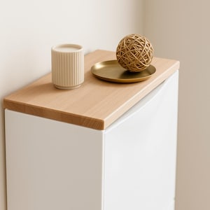 Könnte beinhalten: Eine beige, geriffelte Keramiktasse und ein kleines goldfarbenes Tablett mit einer dekorativen gewebten Kugel stehen auf einer hellen Holzoberfläche. Die Holzoberfläche befindet sich auf einem weißen Schrank. Der Hintergrund ist neutral.