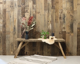 May include: A rustic wooden bench with angled legs, holding a vase of dried flowers and a small plant. A cream-colored rug with gray stripes and fringe lies on the floor. The background is a wooden wall.