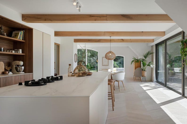 Puede incluir: Interior de cocina moderno y luminoso con encimera blanca y vitrocer&aacute;mica integrada. Vigas y estanter&iacute;as de madera a&ntilde;aden calidez. Se ven una batidora de pie y objetos decorativos. Grandes ventanales ofrecen vistas al exterior.