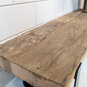 Puede incluir: Un banco rectangular largo con un asiento grueso de madera marr&oacute;n desgastada, que presenta una veta natural prominente y bordes toscos. El asiento est&aacute; sostenido por patas de metal negro, proporcionando una base firme para la superficie.