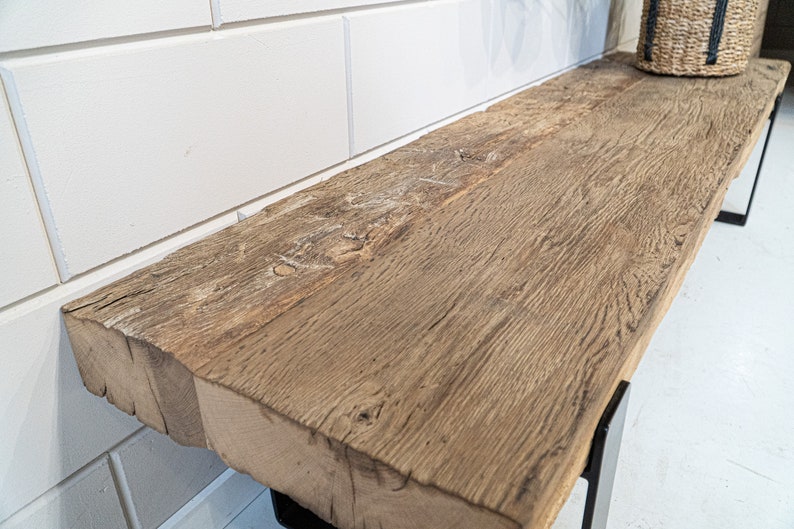 Puede incluir: Un banco rectangular largo con un asiento grueso de madera marr&oacute;n desgastada, que presenta una veta natural prominente y bordes toscos. El asiento est&aacute; sostenido por patas de metal negro, proporcionando una base firme para la superficie.