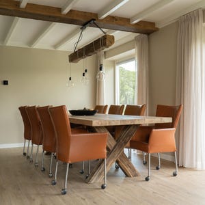 Puede incluir: Una gran mesa de comedor de madera con base en forma de cruz est&aacute; rodeada por diez sillas de cuero marr&oacute;n con ruedas. Una viga de madera con bombillas colgantes est&aacute; suspendida sobre la mesa. La habitaci&oacute;n tiene paredes y suelo de color claro.