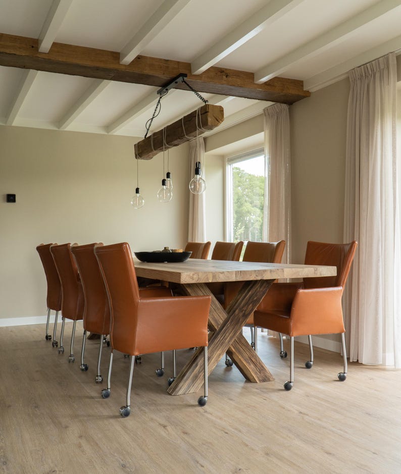 Puede incluir: Una gran mesa de comedor de madera con base en forma de cruz est&aacute; rodeada por diez sillas de cuero marr&oacute;n con ruedas. Una viga de madera con bombillas colgantes est&aacute; suspendida sobre la mesa. La habitaci&oacute;n tiene paredes y suelo de color claro.