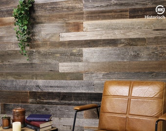 May include: Rustic wood paneling wall with varying shades of brown, gray, and beige. A brown leather armchair with black metal arms is in the foreground. A small table holds books, a candle, and a copper container. Green hanging plant.