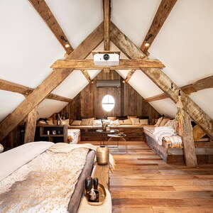 May include: Interior shot of a rustic attic space with exposed wooden beams and a sloped ceiling. The room features a built-in seating area with cushions, a bed, and a projector. The walls are paneled with wood, and the floor is made of wooden planks.