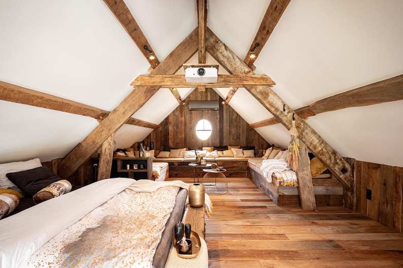 May include: Interior shot of a rustic attic space with exposed wooden beams and a sloped ceiling. The room features a built-in seating area with cushions, a bed, and a projector. The walls are paneled with wood, and the floor is made of wooden planks.